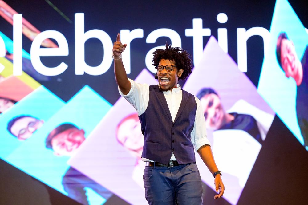 Terrance pointing to the crowd on stage in a waistcoat, a bright screen behind him