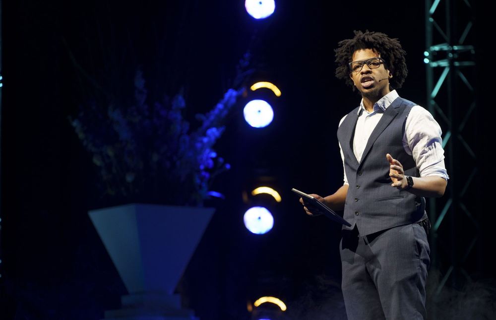 Terrance in a waistcoat gesturing mid-sentence on a dark stage